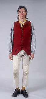 male model wearing long red woolen vest  that button up the front, white patched trousers, a yellow neck scarf and black buckled shoes