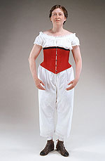 model wearing red corset that hooks up the front, baggy white lace-trimmed drawers that end below the knees and brown and black leather ankle-high boots that tie on the inside of the ankle.